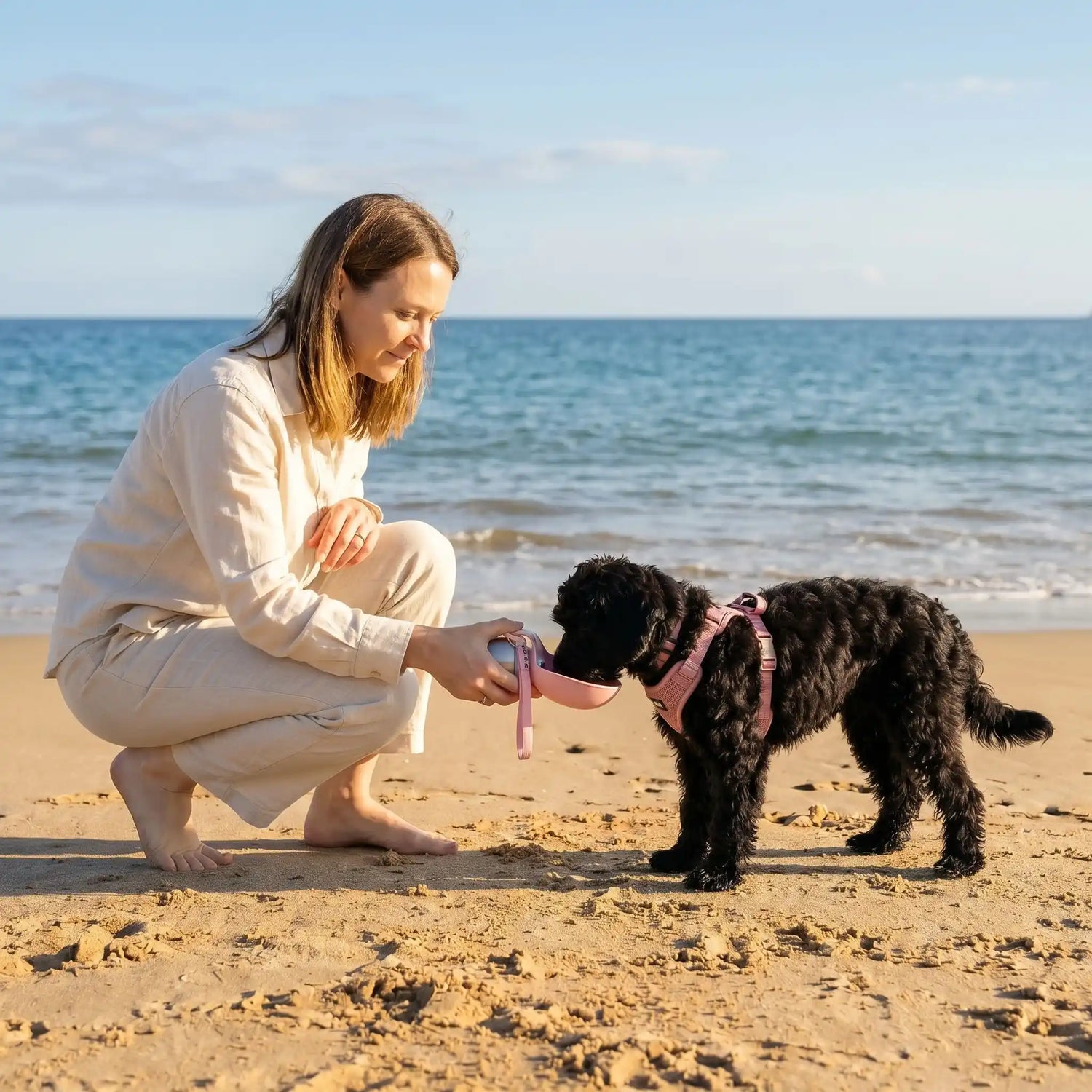 femme qui donne à boire à son chien noir maltipoo avec une gourde rose sur la plage